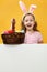 Happy child in rabbit ears holds a basket with Easter eggs on a blank banner space on a yellow background