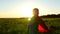 A happy child in a superhero costume in a red raincoat runs along a green lawn against the backdrop of a sunset toward