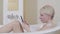Happy confident woman typing on smartphone in bathtub. Side view portrait of blond beautiful Caucasian lady surfing