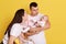 Happy couple embracing and looking at newborn child over yellow background, talking to little daughter with love and smile,