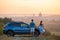 Happy couple relaxing beside their SUV car during honeymoon road trip at sunset. Young man and woman enjoying time together