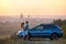 Happy couple spending time together near their SUV car during honeymoon road trip at warm summer evening. Young man and