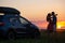 Happy couple standing beside their SUV car during honeymoon road trip at warm summer evening. Young man and woman enjoying time