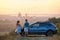 Happy couple standing beside their SUV car during honeymoon road trip at warm summer evening. Young man and woman enjoying time