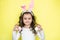 Happy curly little girl wears bunny ears, picks up colored Easter eggs, isolated on yellow background. Easter concept