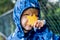 Happy cute caucasian little boy toddler preschooler wearing blue raincoat jacket holding yellow maple leaf on a rainy autumn day.