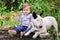 Happy cute female baby and dog is sitting in garden. Child is playing with English Bull Terrier white dog outside in park.