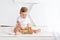 Happy cute little baby six months old in a white t-shirt and diapers sits on a light background at home and plays with a wooden