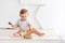 Happy cute little baby six months old in a white t-shirt and diapers sits on a light background at home and plays with a wooden