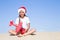 Happy cute little girl wearing a red Santa hat sitting on the sandy beach by the sea and holding a red toy plane on clear sunny da