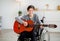 Happy disabled teen boy in wheelchair playing guitar, using musical instrument indoors. Teenage hobbies and pastimes