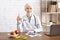 Happy doctor nutritionist looks at camera, holds glass of water, sits at table with apples on plate