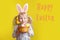 Happy Easter card with text. Caucasian girl in pink  Easter bunny ears holding basket with eggs. Kid child in studio on a yellow