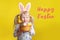 Happy Easter card with text. Caucasian girl in pink  Easter bunny ears holding basket with eggs. Kid child in studio on a yellow