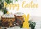 Happy Easter text. Easter greetings lettering. Homemade easter cake, bunny decor, green branches and flowers on wooden background