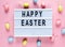 Happy Easter text on lightbox on pink pastel paper background with yellow, pink, blue eggs Bright template for Easter, top view,