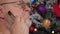 Happy elderly man combing his gray beard with comb on background of Christmas tree in garlands, green UFO balls, proton purple toy