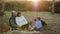 Happy elderly man and woman use computer sitting on plaid with grandson and dog at picnic on background of sunrise