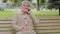 Happy elderly woman with beautiful face sitting on park bench takes phone call, gray-haired lady of retirement age