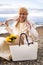Happy elegant blonde woman sitting on pebble sea beach with waving silk scarf and feminine handbag