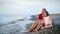 Happy elegant mother and daughter sitting on pebble beach coast together. Wide shot on RED camera