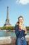 Happy elegant tourist woman with cup of coffee eating croissant