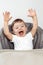 Happy excited adorable baby boy play in high chair with hands up.