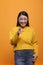 Happy excited beautiful woman wearing yellow sweater pointing index finger at camera on orange background.