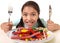 Happy excited Latin female child holding fork and knife sitting at table ready for eat a dish full of candy