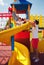 Happy excited toddler baby boy sliding in colorful playground at summer day with the help of father