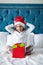 Happy exited little girl in Santa`s hat holding gift box, looking at camera excited while sitting on bed