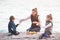 Happy family mother and two kids in spring clothes drink hot tea on windy cold sea beach during leisure picnic