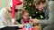 Happy family, parents showing Ney Year decorations to their cute child in front of Christmas tree. Mother in Santa hat