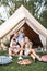 Happy family on picnic, sitting on green grass near the big white tipi tent in forest or park. Father, mother and two