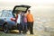 Happy family standing together near a car with open trunk enjoying view of rural landscape nature. Parents and their kids leaning