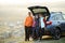 Happy family standing together near a car with open trunk enjoying view of rural landscape nature. Parents and their kids leaning