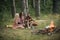 Happy friends enjoying picnic in woods. Bearded man and two sisters in retro dresses sitting next to campfire and