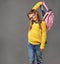 Happy fun positive school girl holding pink color backpack above the head in yellow sweater and blue jeans on grey background.