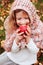 Happy funny kid girl in cozy knitted scarf eating fresh apple in autumn garden