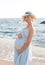 Happy future mother hug her belly, calm pregnant woman dressed in blue dress and hat have a rest on sea beach