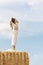 Happy girl enjoys fresh air in the countryside. Young beautiful woman with long hair stands on haystack against blue sky