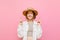 Happy girl in light clothing and hat stands on a pink background and makes a wish with crossed fingers and buried eyes, smiling.