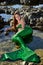 A happy girl in a mermaid costume lies in the water among the stones on the beach and looks at the mermaid tail.