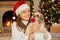 Happy girl in santa hat and white sweater hugging with cute Pekingese dog on background of beautiful christmas tree with lights