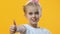 Happy girl showing thumbs-up standing on yellow background, children recommend