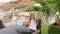 Happy girl sitting under palm tree leaves on outdoor balcony with sea view. Smiling girl on summer balcony in resort