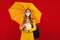 Happy girl student in a medical protective mask on her face, with a backpack and books, and a yellow umbrella in her hands stands