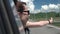 Happy girl in sunglasses leaning out of car window and enjoying trip in mountains. Young woman looking out window of