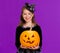 Happy girl in witch costume with pumpkin jack-o-lantern celebrates Halloween and laughs on violet background