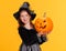 Happy girl in witch costume with pumpkin jack-o-lantern celebrates Halloween and laughs on yellow background